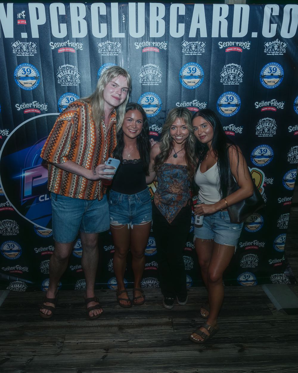 Four friends posing in front of the PCB Club Card photo wall at Hammerhead Fred's